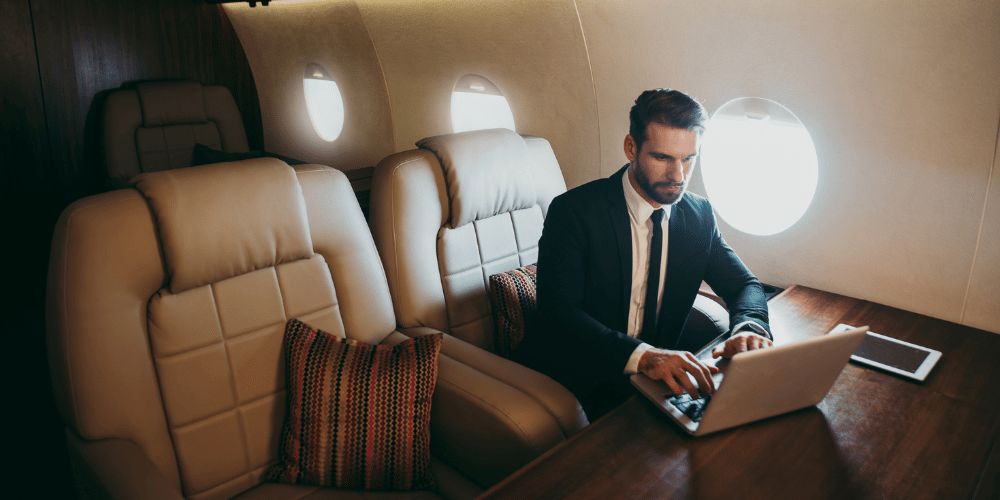 Ultra-high net-wroth businessman on private jet looking at his private client reputation strategy on a laptop - The PHA Group